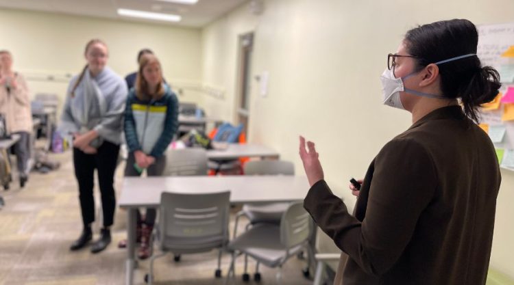 Women wearing mask (Marie Spiker) addresses students in a class who are gathered around for a group activity